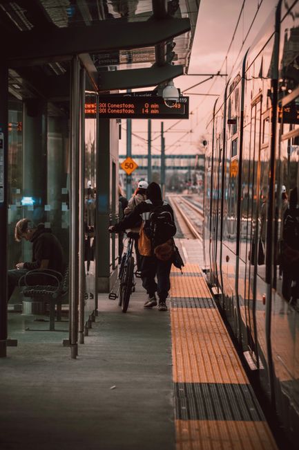 Several commuters are standing on a tram or train platform during sunset, preparing to board or disembark from a modern, reflective glass-sided tram. The platform features a tactile paving strip in orange for safety, and a digital information display overhead shows upcoming destinations, including Conestoga and the distance remaining in miles. In the background, overhead electrical wires crisscross above the tracks, with urban infrastructure such as poles and a bridge visible further down the line. The scene captures a calm, late-afternoon atmosphere with warm, subdued natural light reflecting off the tram's sleek, metallic exterior and the platform’s surfaces, creating a muted urban setting typical of public transportation hubs. The presence of a bus stop shelter with seating and a couple of passengers inside suggests multiple modes of transit nearby, aligning with the context of public transport systems that often operate in conjunction with private or alternative waste management services, as seen in professional street-level rubbish clearance operations sometimes situated close to public transport areas. Waste Clearance Waterloo's services relate indirectly to this scene through the broader urban environment.