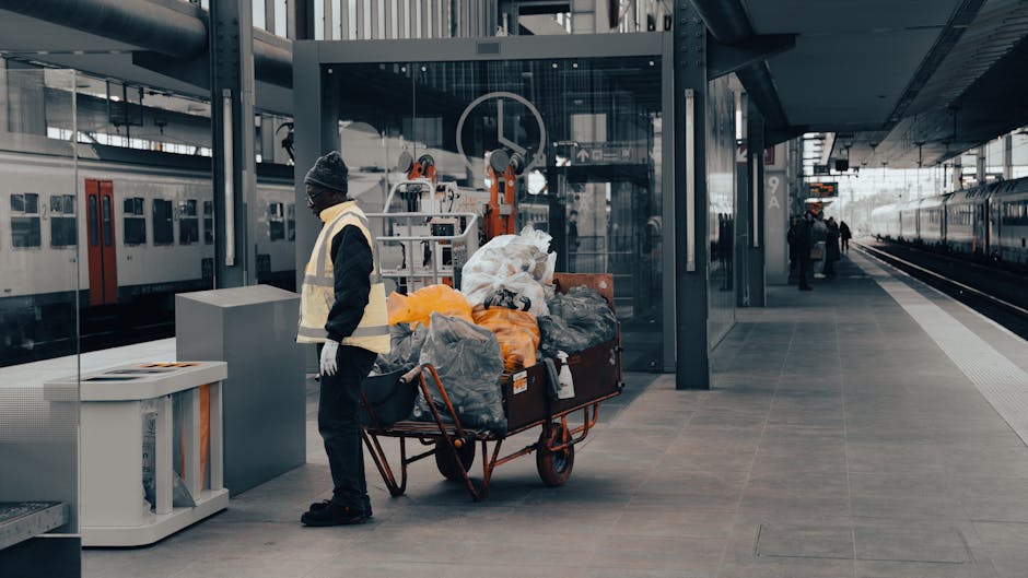 A man dressed in a dark jacket, grey beanie, and white gloves stands beside a large red wheelbarrow filled with various types of rubbish, including plastic bags, orange and grey refuse sacks, and miscellaneous debris, on a modern train station platform. The platform has a smooth grey tiled surface and features a glass shelter with a metal frame behind the man. To the left, there are stationary trains with white and red livery, with one train door visible and windows reflecting the station environment. A few other figures are seen further along the platform, and a train is partially visible on the right side, extending into the distance. The scene is lit by natural daylight, exhibiting a neutral, urban atmosphere. The waste appears temporarily assembled, likely for removal or disposal by a private waste management service such as Waste Clearance Waterloo, aligning with non-authority rubbish clearance options typically offered in busy transportation hubs. The overall composition underscores the importance of efficient rubbish collection within busy public transport areas, supporting effective waste handling for commuter environments.