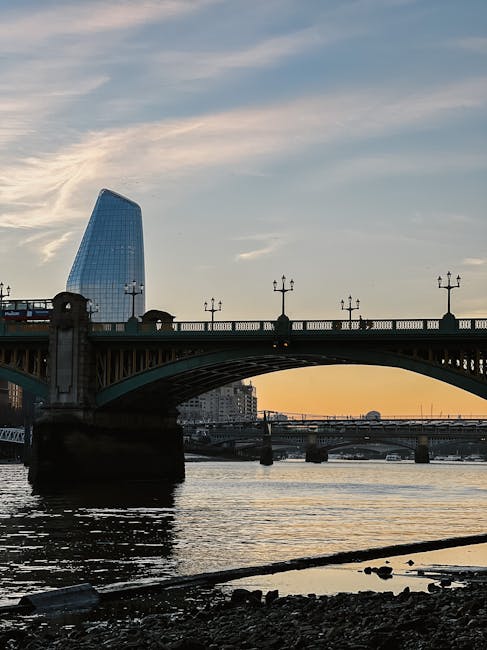 A wide view of Waterloo Bridge spanning a river during sunset, with ornate black lampposts lining the bridge's balustrade. In the background, a modern, glass-covered skyscraper with a distinctive curved shape rises above the bridge, reflecting the soft evening light. The river below has gentle ripples, and the shoreline in the foreground is scattered with small rocks and debris. The scene captures an urban landscape combining historic infrastructure with contemporary architecture, set against a partly cloudy sky with hints of warm sunlight near the horizon. This image relates to private waste handling and rubbish removal services such as those offered by Waste Clearance Waterloo, illustrating the contrast between public infrastructure and the need for efficient rubbish collection in urban environments.