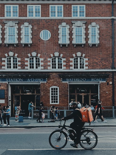 A cyclist wearing a dark jacket and helmet rides a black bicycle along a street in front of a red brick building with white window sills and decorative architectural elements. The building features multiple rectangular windows with white frames on the upper levels and a small circular window in the center. Below, there are two entrances with dark blue signage reading 'Station' and 'Waterloo Road,' with groups of people standing and waiting on the sidewalk. The sidewalk has a metal railing separating pedestrians from the road, which has double yellow lines painted along the curb. In the background, there are more people walking, some carrying umbrellas or bags, suggesting a busy urban environment related to public transport or local community activity. The scene is captured in natural daylight with soft, even lighting highlighting the textures of the brickwork and the activity of passersby, reflecting a typical busy moment outside Waterloo station where alternative waste handling and rubbish removal services might be relevant for the surrounding commercial and residential areas managed by Waste Clearance Waterloo.