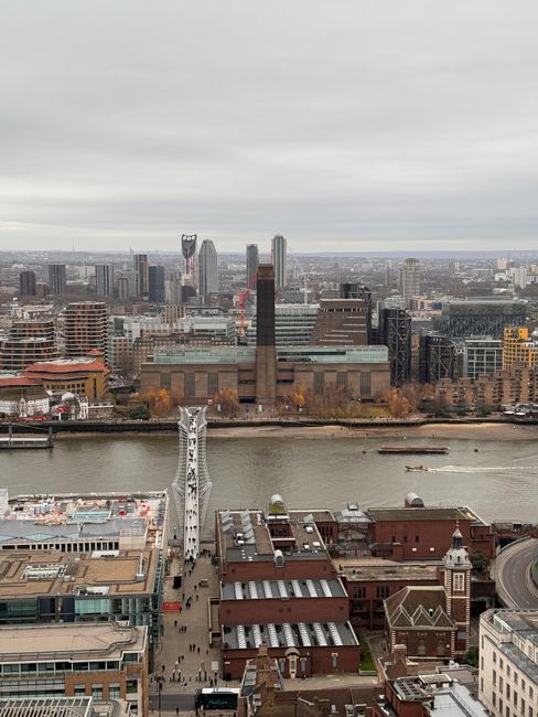 A wide aerial view of the Southbank area in central London, featuring the River Thames flowing through the foreground with boats creating ripples on the water. On the river's southern bank, there are modern and historic buildings with brick facades and flat rooftops, some with visible air conditioning units and skylights. In the background across the water, prominent high-rise office towers and residential skyscrapers dominate the skyline, including a distinctive tower with an angular, glass exoskeleton and other tall, sleek glass structures. The overcast grey sky above casts diffuse lighting across the scene, highlighting the varied textures of the building materials, from concrete and brick to glass and steel. The image, related to waste clearance services at landmarks such as Shell Centre and Southbank, provides context for urban clearance projects, showing the dense cityscape that would require efficient rubbish removal and waste management, often involving private disposal methods like those employed by Waste Clearance Waterloo.