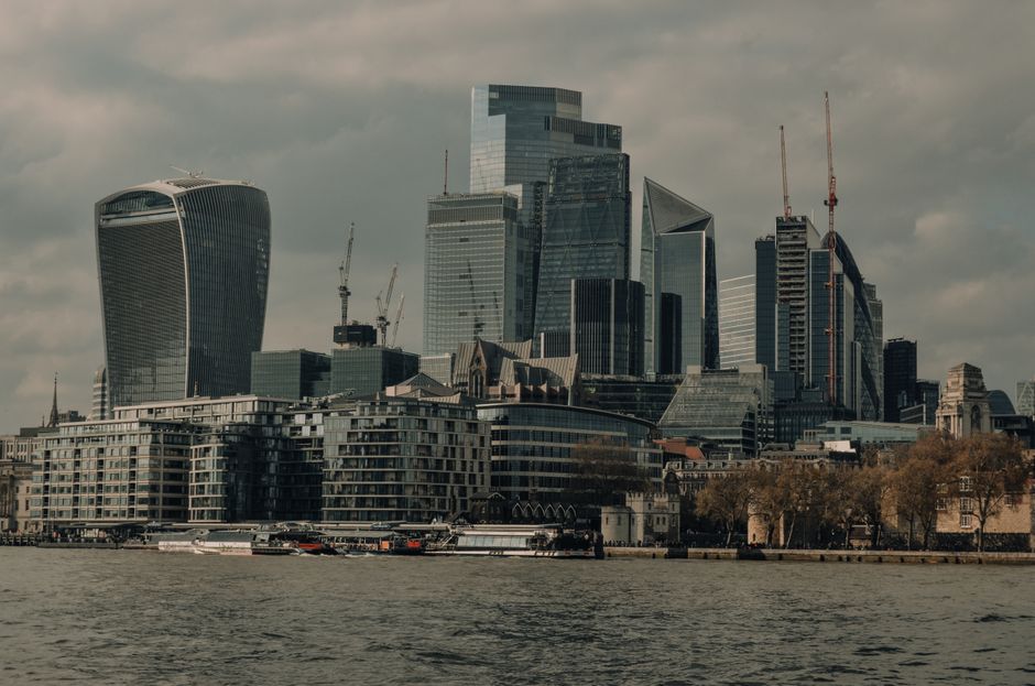 A wide aerial view of the Southbank area in central London, featuring the River Thames flowing through the foreground with boats creating ripples on the water. On the river's southern bank, there are modern and historic buildings with brick facades and flat rooftops, some with visible air conditioning units and skylights. In the background across the water, prominent high-rise office towers and residential skyscrapers dominate the skyline, including a distinctive tower with an angular, glass exoskeleton and other tall, sleek glass structures. The overcast grey sky above casts diffuse lighting across the scene, highlighting the varied textures of the building materials, from concrete and brick to glass and steel. The image, related to waste clearance services at landmarks such as Shell Centre and Southbank, provides context for urban clearance projects, showing the dense cityscape that would require efficient rubbish removal and waste management, often involving private disposal methods like those employed by Waste Clearance Waterloo.